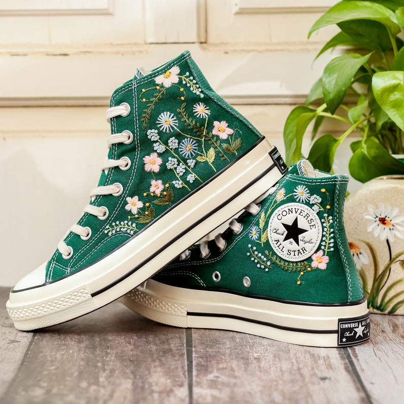 Puede incluir: Zapatillas altas verde esmeralda con bordados florales. Las zapatillas tienen cordones blancos, puntera de goma blanca y suela color crema. El logo de Converse All Star es visible en el lateral.