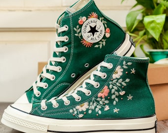 May include: Green high-top Converse sneakers with white soles and floral embroidery. The Converse logo is embroidered on the side of the shoe.