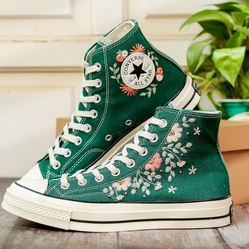 May include: Green high-top Converse sneakers with white soles and floral embroidery. The Converse logo is embroidered on the side of the shoe.