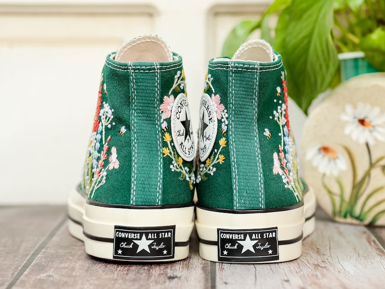 Może przedstawiać: Para zielonych, wysokich trampek Converse Chuck Taylor z haftem kwiatowym. Trampki mają białą gumową podeszwę i czarne szwy.
