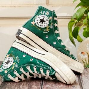 Puede incluir: Zapatillas altas verdes con bordados florales. Las zapatillas tienen cordones blancos, suelas y un parche con el logo de la estrella negra que dice "Converse All Star". Los zapatos son de lona y tienen un dise&ntilde;o cl&aacute;sico.