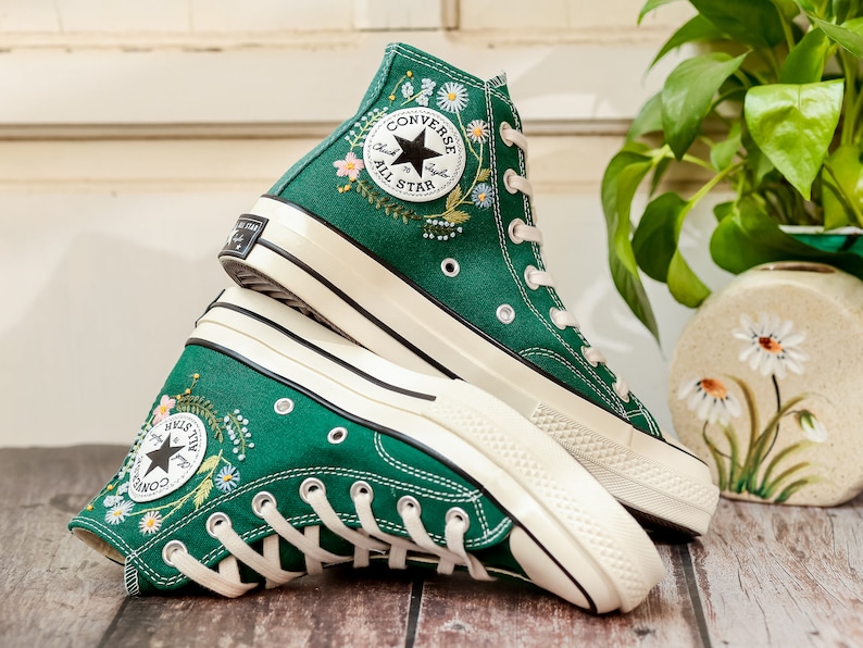 Puede incluir: Zapatillas altas verdes con bordados florales. Las zapatillas tienen cordones blancos, suelas y un parche con el logo de la estrella negra que dice "Converse All Star". Los zapatos son de lona y tienen un dise&ntilde;o cl&aacute;sico.
