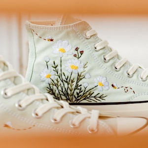 May include: A pair of light green canvas high-top sneakers with white laces and embroidered white daisies and green leaves. The sneakers have a white rubber sole.