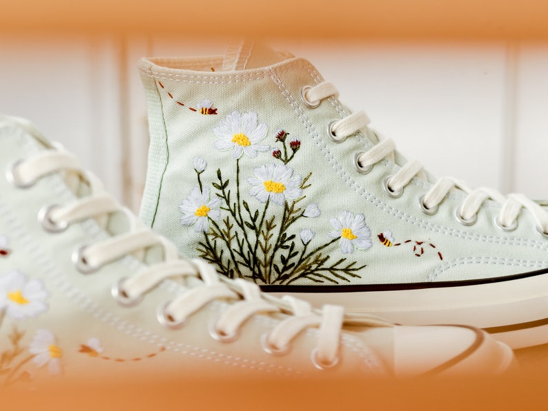 May include: A pair of light green canvas high-top sneakers with white laces and embroidered white daisies and green leaves. The sneakers have a white rubber sole.