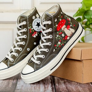 May include: A pair of olive green high-top Converse trainers with white soles and red and white embroidered flowers and mushrooms. The trainers are on a brown cardboard box.