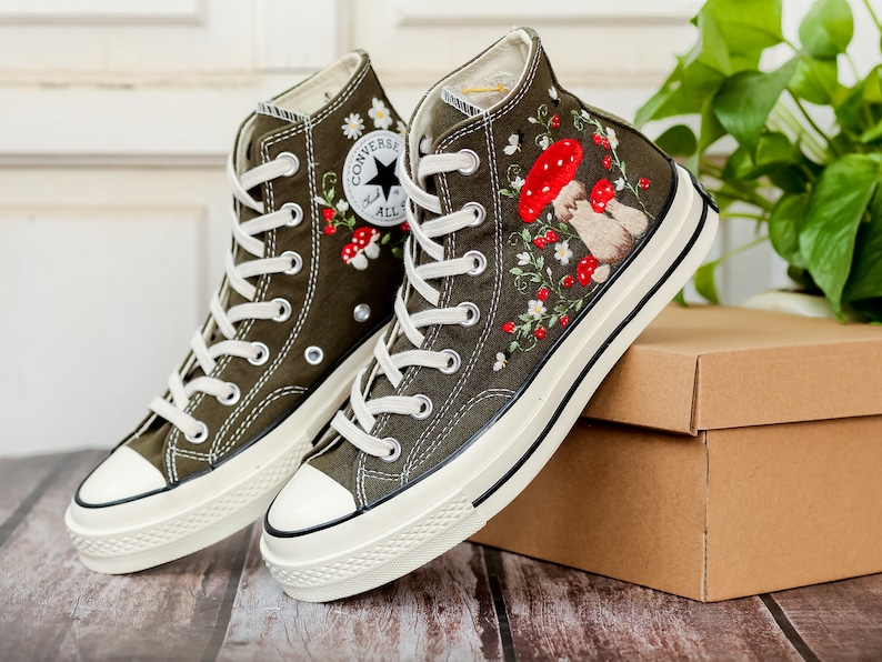 May include: A pair of olive green high-top Converse trainers with white soles and red and white embroidered flowers and mushrooms. The trainers are on a brown cardboard box.