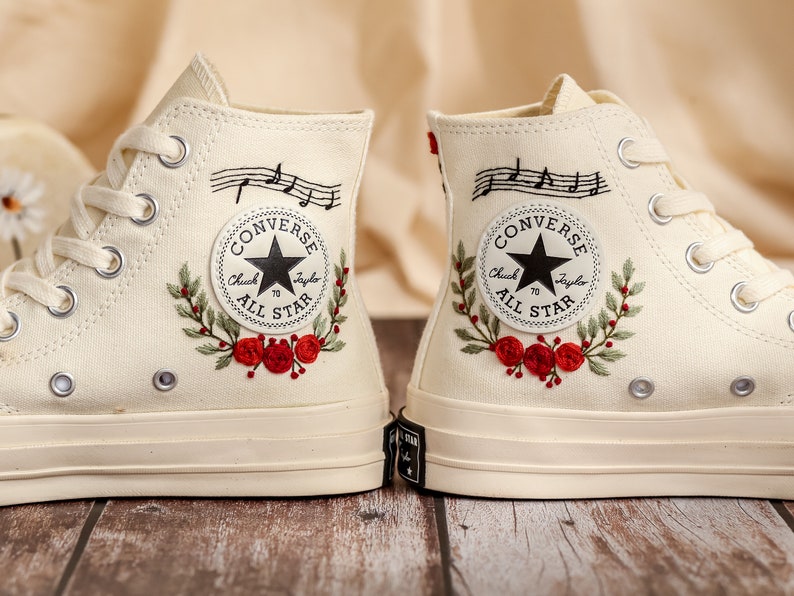 May include: A pair of off-white high-top Converse sneakers with red embroidered flowers and musical notes on the side. The Converse logo is black with a white star and the words "Chuck Taylor All Star".