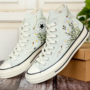 May include: A pair of light blue high-top canvas sneakers with white laces and a white rubber sole. The sneakers are decorated with embroidered white flowers and green leaves. The Converse All Star logo is visible on the side of the sneakers.