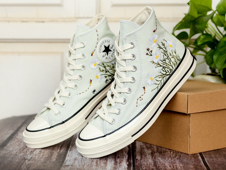 May include: A pair of light blue high-top canvas sneakers with white laces and a white rubber sole. The sneakers are decorated with embroidered white flowers and green leaves. The Converse All Star logo is visible on the side of the sneakers.
