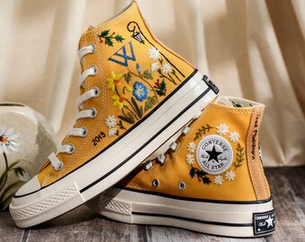 May include: Mustard yellow high-top sneakers with white laces and a cream-colored sole. The sneakers feature embroidered floral designs, the word "2019", and the Converse All Star logo. The shoes are on a wooden surface.