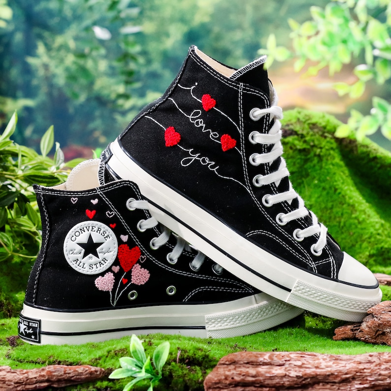 Op de afbeelding: Zwarte Converse sneakers met hoge schacht en witte zolen. De ene schoen heeft rode geborduurde hartjes en de tekst "I love you". De andere schoen heeft rode en roze geborduurde hartjes met een Converse All Star logo.