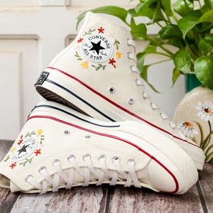 May include: A pair of white high-top Converse trainers with red trim and embroidered flowers. The trainers have a black star logo and the text "Converse All Star" on the side.