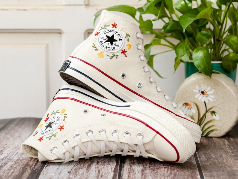 May include: A pair of white high-top Converse trainers with red trim and embroidered flowers. The trainers have a black star logo and the text "Converse All Star" on the side.