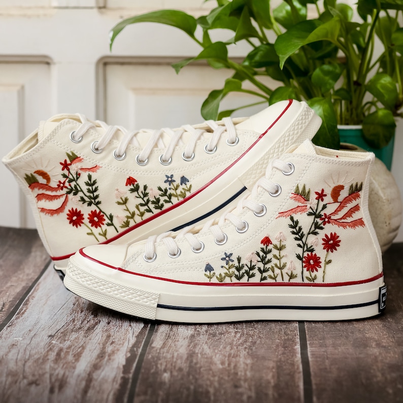 May include: A pair of white high-top canvas trainers with embroidered floral designs in shades of red, pink, green, and blue. The trainers have a white rubber sole.