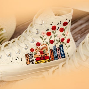 May include: White high-top canvas sneakers with embroidered flowers, musical notes, and a stack of books with titles like "Speak Now", "Fearless", "Red", "1989", "Lover", "Evermore", and "Midnights".