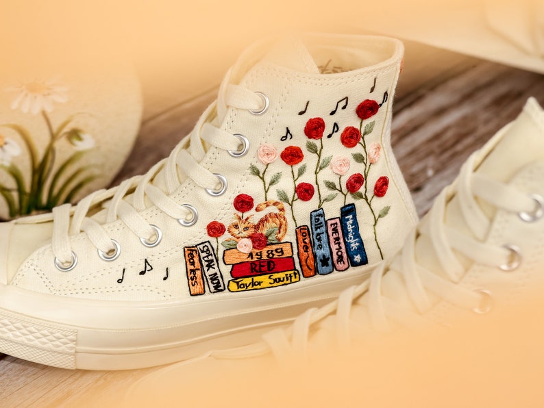 May include: White high-top canvas sneakers with embroidered flowers, musical notes, and a stack of books with titles like "Speak Now", "Fearless", "Red", "1989", "Lover", "Evermore", and "Midnights".