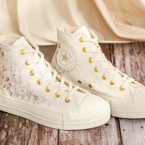 May include: A pair of off-white high-top trainers with gold eyelets and embroidered floral details. The trainers have a white rubber sole and a white canvas upper. The Converse All Star logo is visible on the side of the trainers.