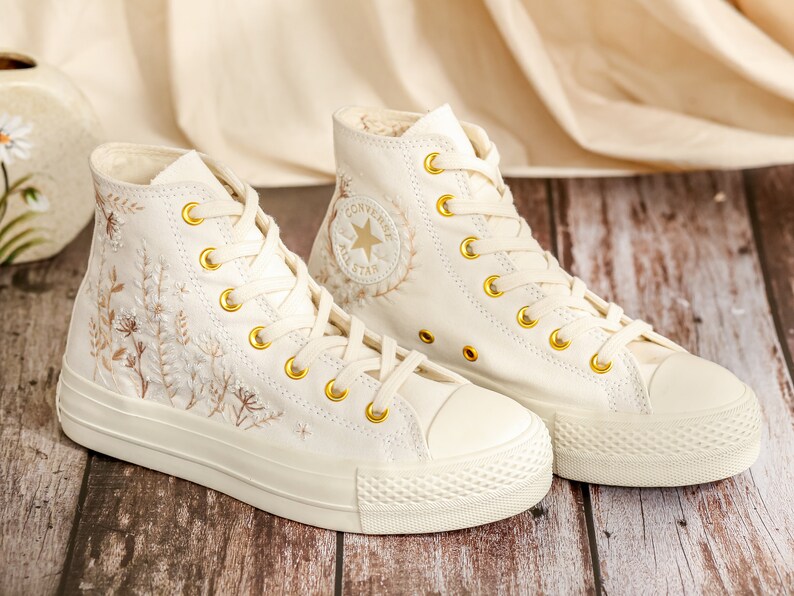 May include: A pair of off-white high-top trainers with gold eyelets and embroidered floral details. The trainers have a white rubber sole and a white canvas upper. The Converse All Star logo is visible on the side of the trainers.