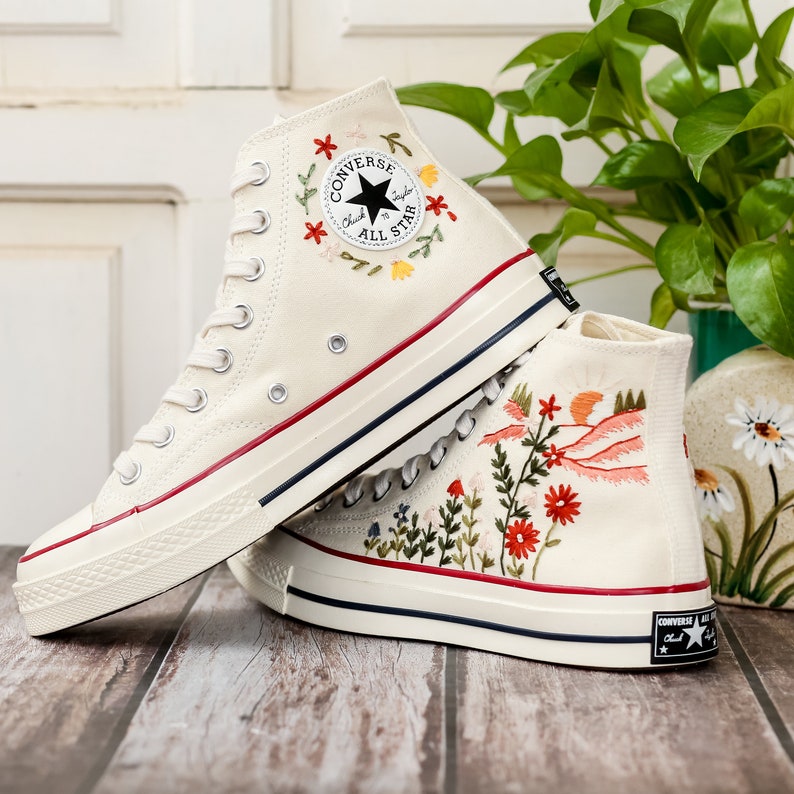May include: A pair of white high-top Converse trainers with embroidered floral designs. One shoe features a floral design around the Converse All Star logo, while the other shoe has a more elaborate floral design with red, pink, and orange accents.