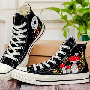 May include: A pair of black high-top canvas sneakers with white soles and laces. The sneakers are decorated with embroidered red mushrooms, green leaves, and yellow flowers.