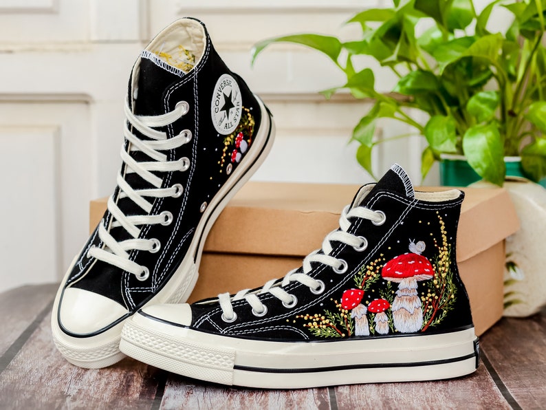 May include: A pair of black high-top canvas sneakers with white soles and laces. The sneakers are decorated with embroidered red mushrooms, green leaves, and yellow flowers.