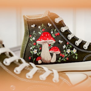 May include: A pair of green high-top canvas shoes with white laces and embroidered red and white mushrooms and flowers.