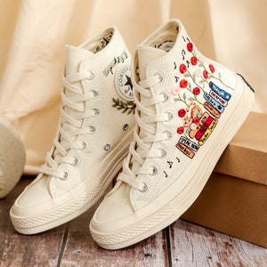 May include: A pair of off-white high-top canvas sneakers with embroidered flowers and a stack of books on one shoe. The other shoe has embroidered musical notes and a small green plant.