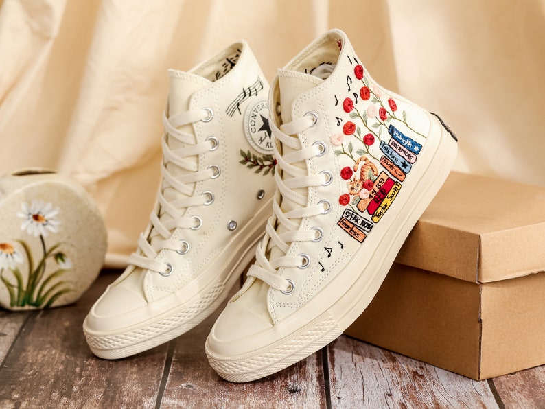 May include: A pair of off-white high-top canvas sneakers with embroidered flowers and a stack of books on one shoe. The other shoe has embroidered musical notes and a small green plant.