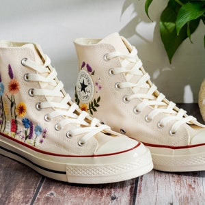 K&ouml;nnte beinhalten: Ein Paar cremefarbene High-Top-Canvas-Sneaker mit gestickten Blumenmustern. Die Sneaker haben wei&szlig;e Schn&uuml;rsenkel und einen roten Rand um die Sohle.