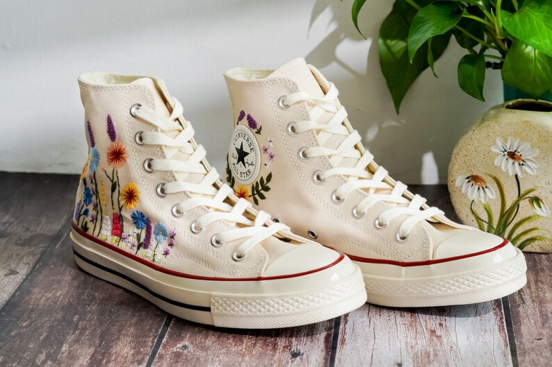 K&ouml;nnte beinhalten: Ein Paar cremefarbene High-Top-Canvas-Sneaker mit gestickten Blumenmustern. Die Sneaker haben wei&szlig;e Schn&uuml;rsenkel und einen roten Rand um die Sohle.
