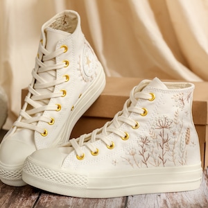 May include: A pair of off-white high-top canvas trainers with gold eyelets and embroidered floral details. The trainers are on a wooden surface.