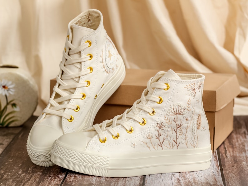 May include: A pair of off-white high-top canvas trainers with gold eyelets and embroidered floral details. The trainers are on a wooden surface.