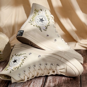 May include: A pair of white high-top Converse sneakers with floral embroidery and the Converse All Star logo. The sneakers are on a wooden surface.