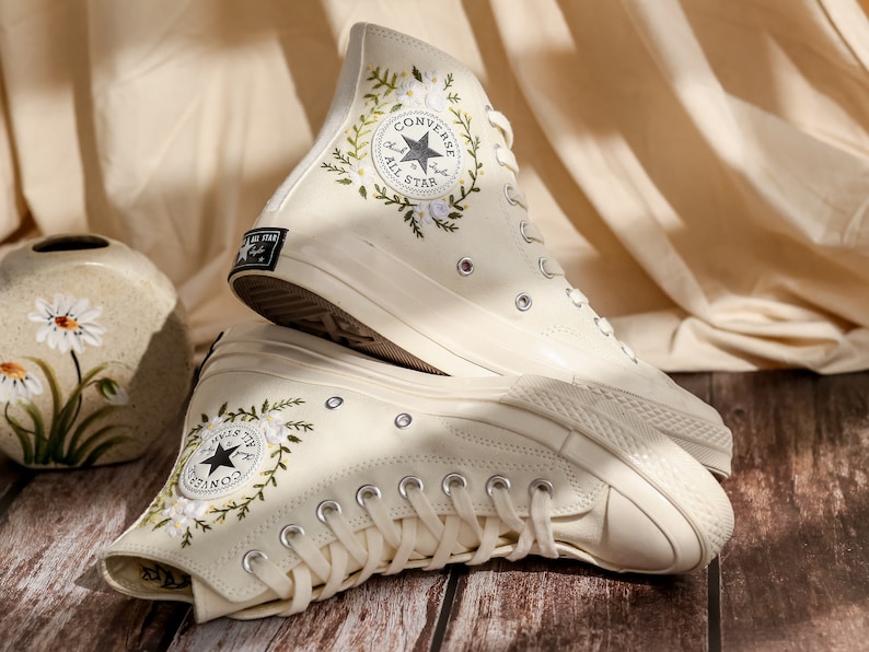 May include: A pair of white high-top Converse sneakers with floral embroidery and the Converse All Star logo. The sneakers are on a wooden surface.