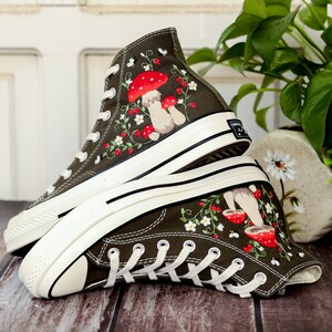 May include: A pair of dark green high-top canvas trainers with white soles. The trainers are decorated with embroidered red mushrooms, white flowers, and green leaves.