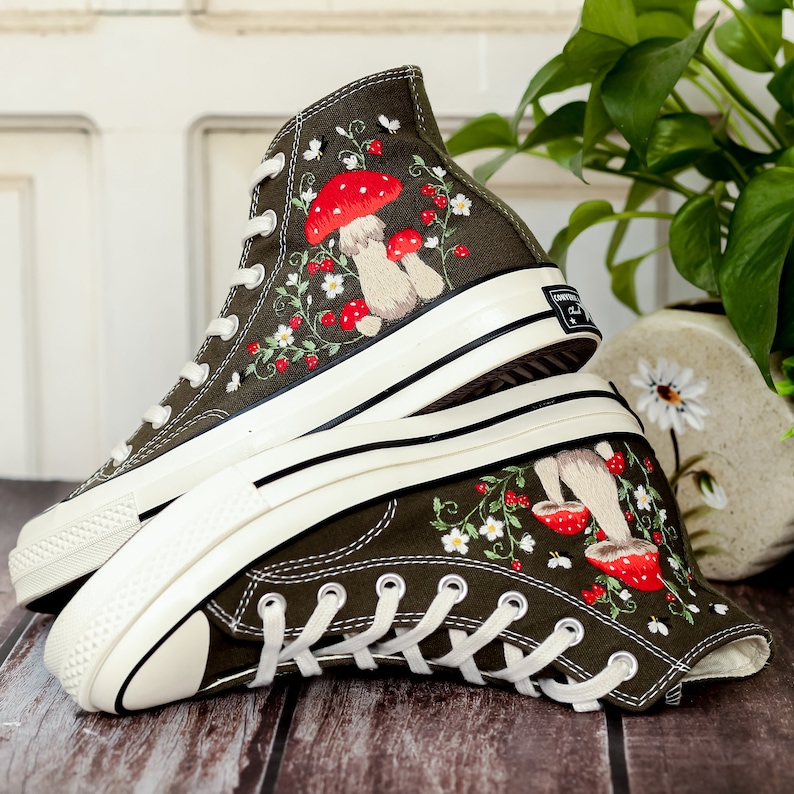 May include: A pair of dark green high-top canvas trainers with white soles. The trainers are decorated with embroidered red mushrooms, white flowers, and green leaves.