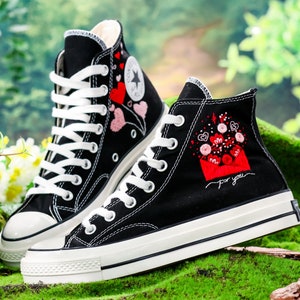 Op de afbeelding: Een paar zwarte high-top canvas sneakers met witte veters en een witte rubberen zool. De sneakers zijn geborduurd met rode en roze harten en de tekst "For you" aan de zijkant.