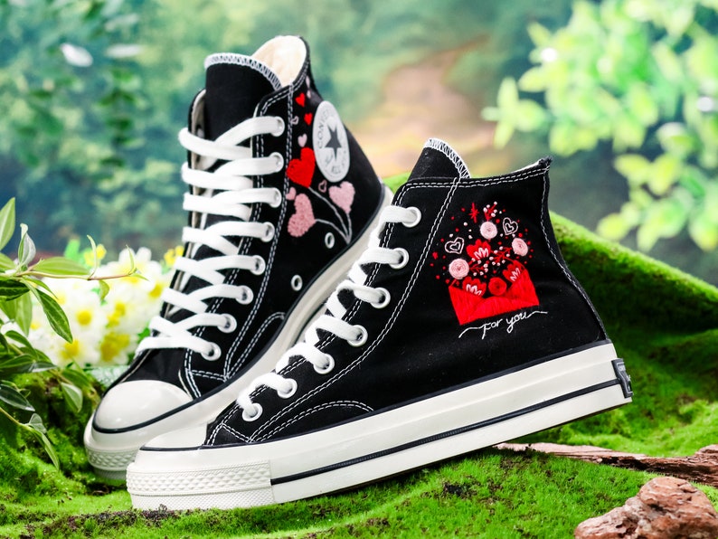 Op de afbeelding: Een paar zwarte high-top canvas sneakers met witte veters en een witte rubberen zool. De sneakers zijn geborduurd met rode en roze harten en de tekst "For you" aan de zijkant.