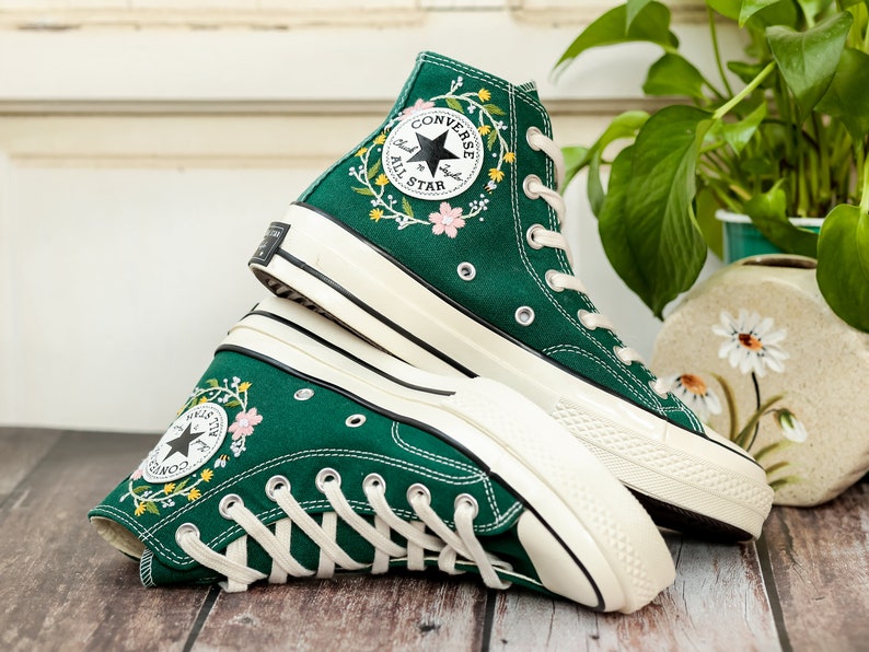 Może przedstawiać: Para zielonych, wysokich trampek Converse z białymi podeszwami i haftem kwiatowym. Logo Converse jest widoczne z boku but&oacute;w.