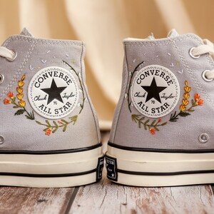 May include: A pair of gray high-top Converse sneakers with embroidered flowers and the text "Converse Chuck Taylor All Star" on the side.