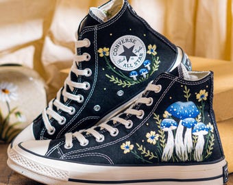May include: Black high-top sneakers with white laces and soles. The sneakers feature embroidered blue mushrooms and yellow flowers. The Converse logo is visible on the side. The shoes are on a wooden surface.
