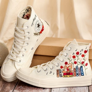 May include: A pair of white high-top canvas sneakers with embroidered floral designs and music notes. One shoe features a stack of books with the text "Taylor Swift" and album titles. The other shoe has a red Converse logo.