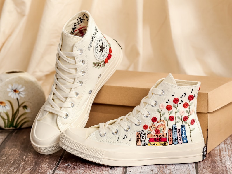 May include: A pair of white high-top canvas sneakers with embroidered floral designs and music notes. One shoe features a stack of books with the text "Taylor Swift" and album titles. The other shoe has a red Converse logo.