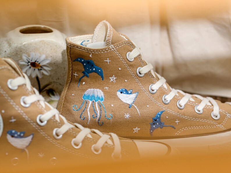 May include: A pair of beige canvas high-top sneakers with embroidered ocean-themed designs, including jellyfish, manta rays, and stars.