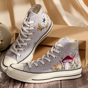 May include: A pair of gray high-top Converse sneakers with embroidered floral and mushroom designs. The sneakers have white soles and laces.