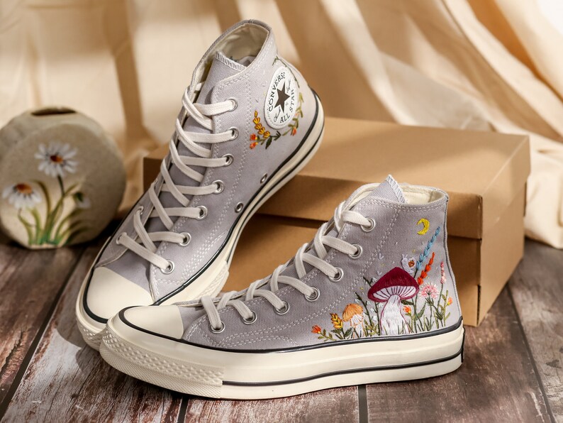 May include: A pair of gray high-top Converse sneakers with embroidered floral and mushroom designs. The sneakers have white soles and laces.