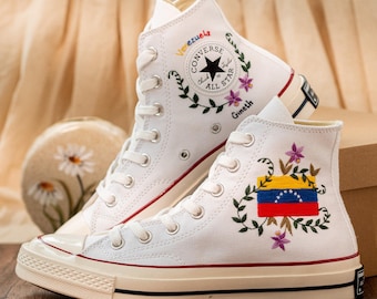Può includere: Un paio di sneakers alte bianche con dettagli rossi e crema. Le sneakers presentano ricami floreali e la bandiera venezuelana. Il logo Converse All Star è visibile, con la parola "Venezuela" sopra.