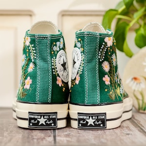 Puede incluir: Zapatillas altas de lona verde esmeralda, adornadas con dise&ntilde;os florales bordados en blanco, rosa y azul. Presentan el logo Converse All Star en el tobillo y suela blanca.