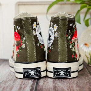 May include: A pair of green canvas high-top trainers with embroidered flowers and mushrooms. The trainers have white soles and black laces. The text "Converse All Star Chuck Taylor" is printed on the back of the trainers.