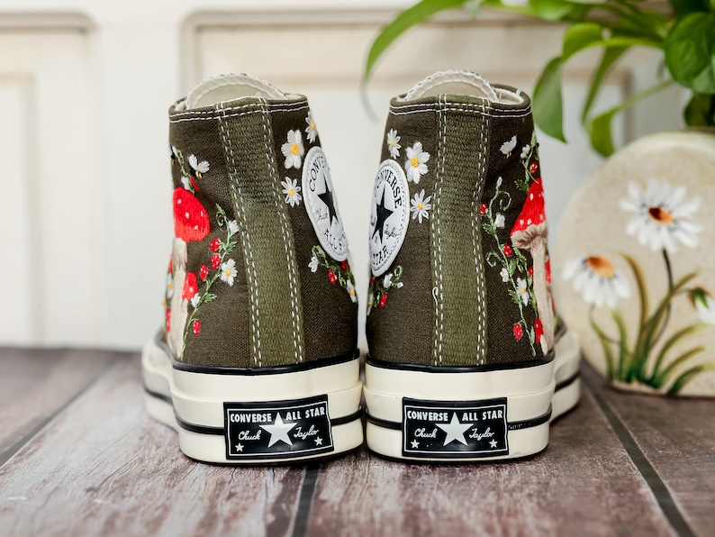 May include: A pair of green canvas high-top trainers with embroidered flowers and mushrooms. The trainers have white soles and black laces. The text "Converse All Star Chuck Taylor" is printed on the back of the trainers.